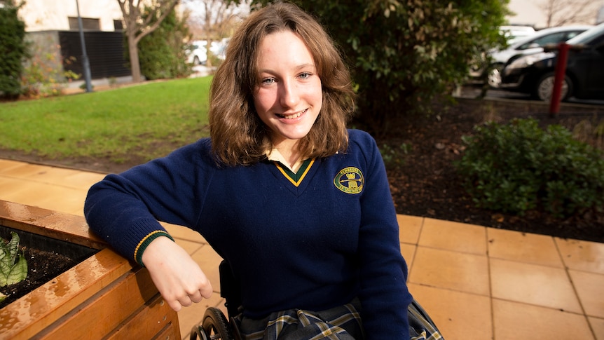 A red-haired schoolgirl in a navy jumper with her school's logo on it, sitting in her wheelchair, smiling at the camera