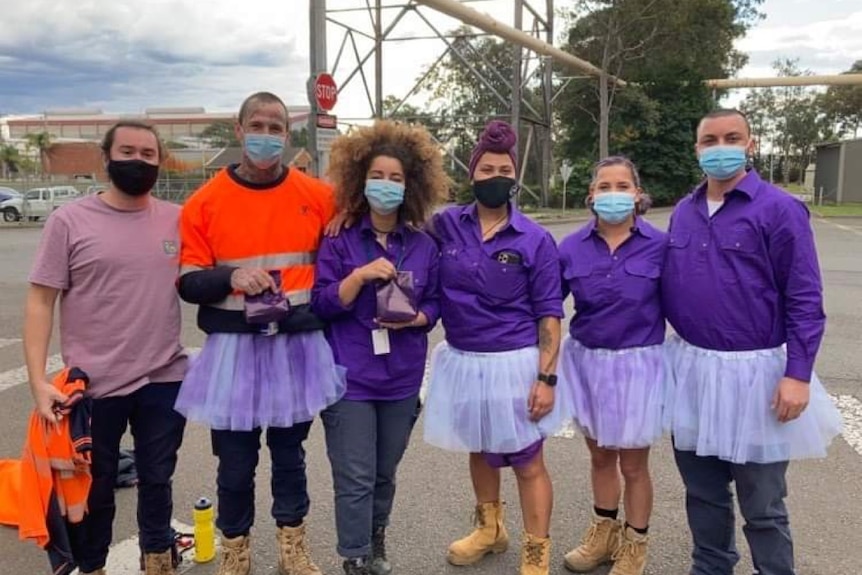A group of people stand wearing purple shirts, tutus, and orange hi vis at a grain handling site.