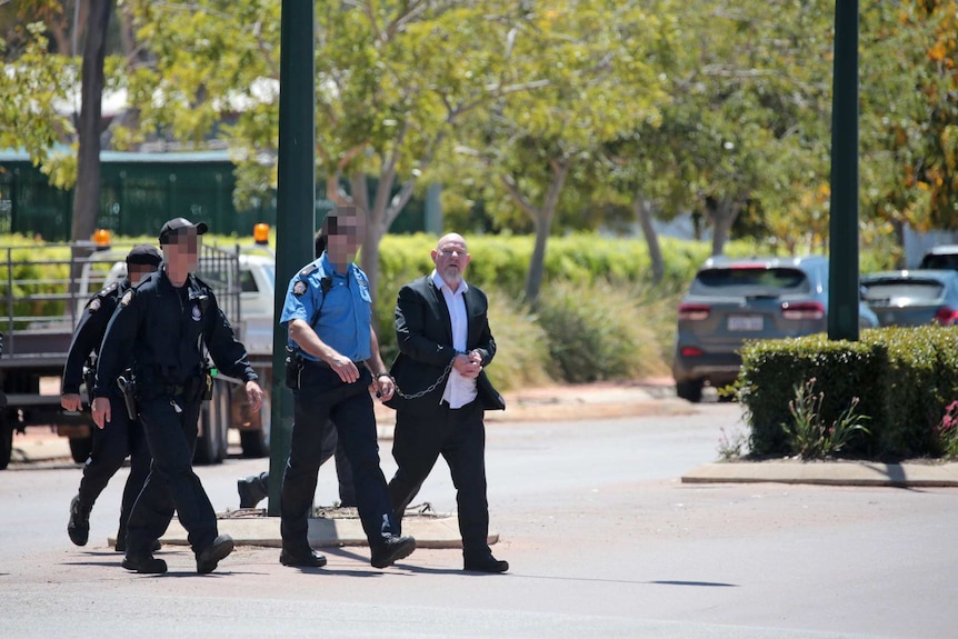 Wark walks across a road with four police officers, one of whom he is chained to. He wears a black suit and white shirt.