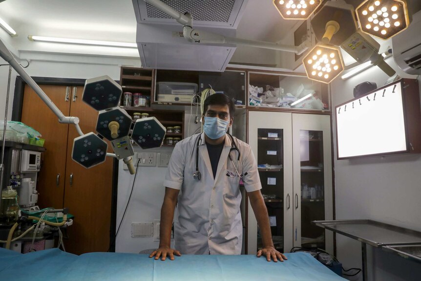 An Indian doctor in a face mask and scrubs standing over an empty hospital bed