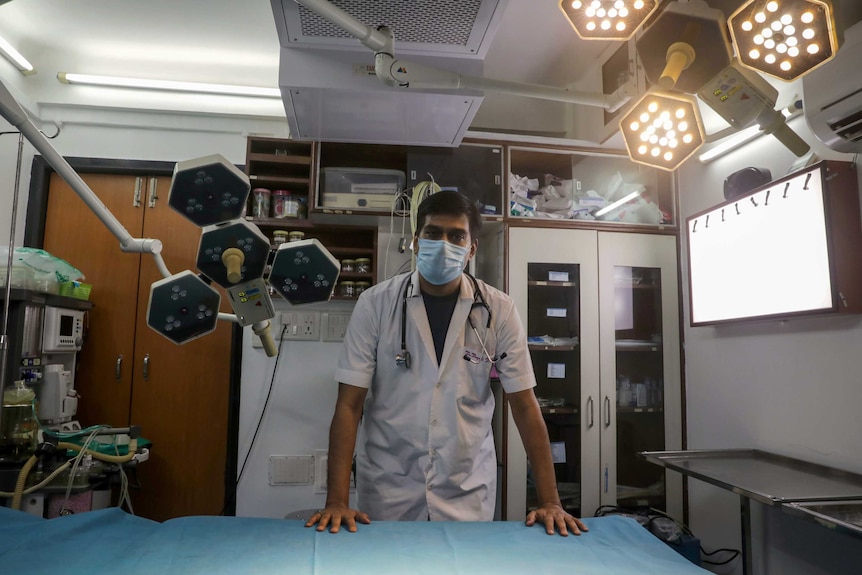 An Indian doctor in a face mask and scrubs standing over an empty hospital bed