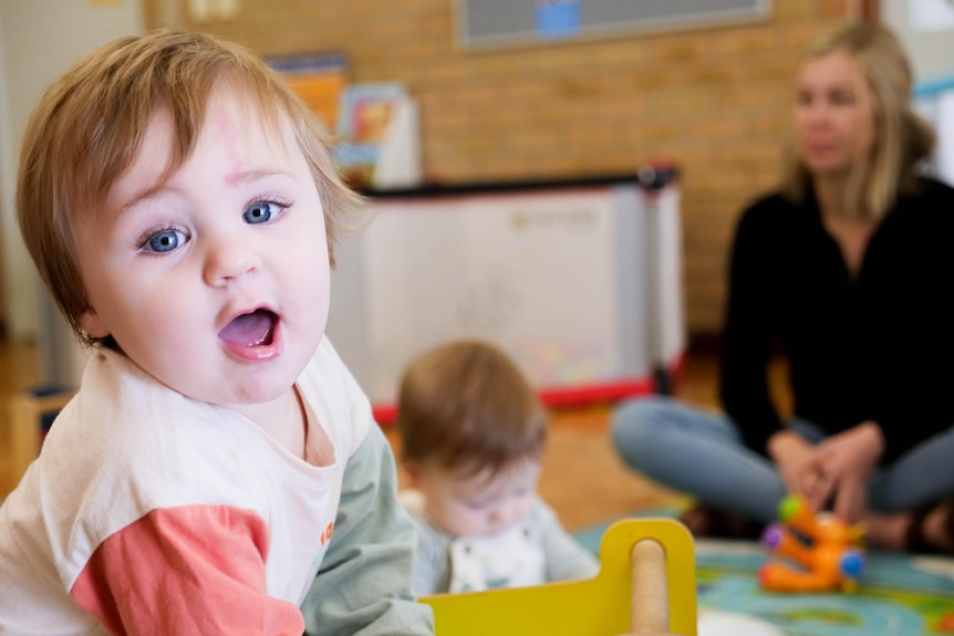 A baby boy with an open mouth half smiling in the foreground and another baby and adult in the background