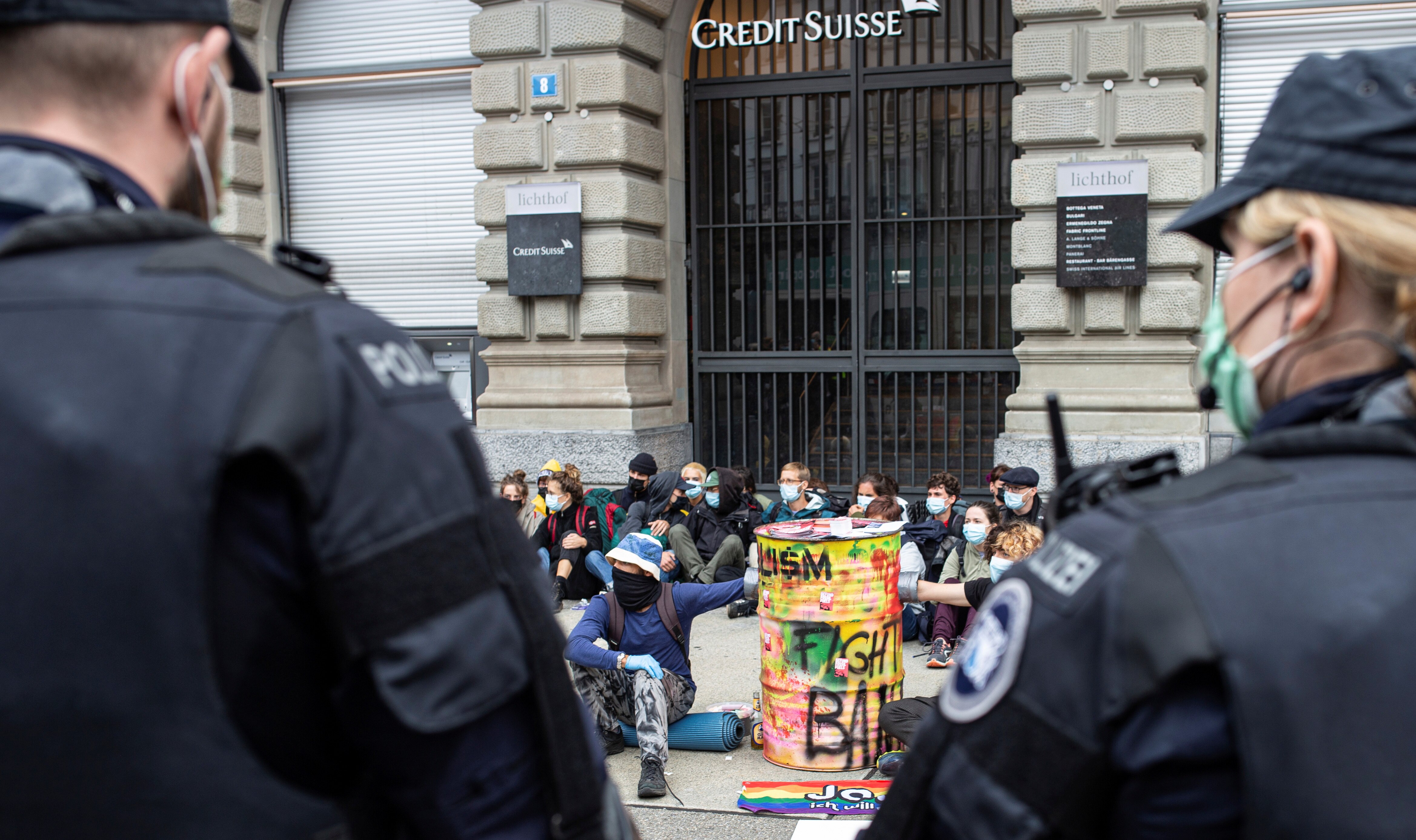 A male and female police officer in the foreground prepare to approach a group of people who sit on ground in front of a bank.