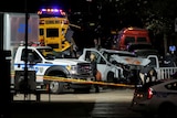 A ute with a smashed bonnet sits behind police tape in a New York street at night time, with police vehicles and a school bus.
