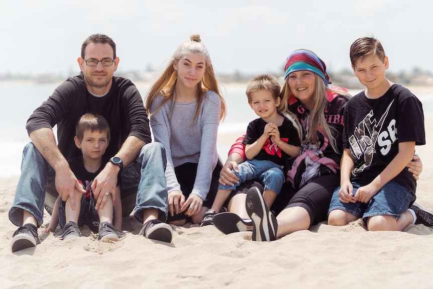 A family including mum and dad and four children, three boys and one girl, sit on a beach posing for a photo.