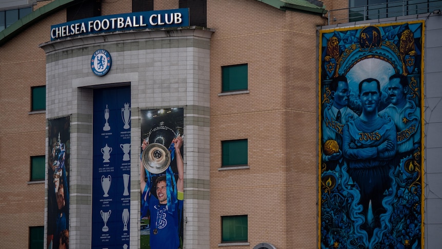 A sign above a stadium says "Chelsea Football Club", with pictures next to it of Chelsea players lifting trophies.