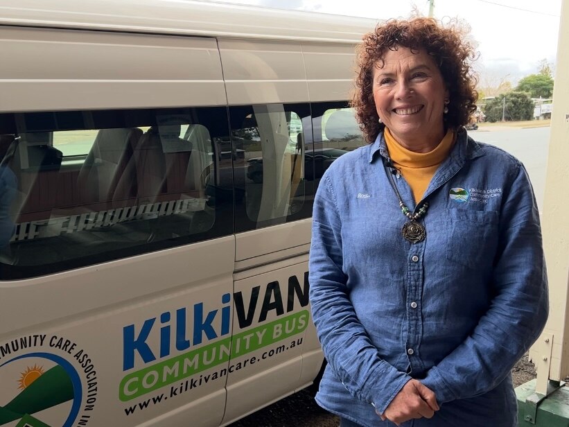 A lady with curly red hair stands on the side of the road smiling in front of a white mini-van, with KilkiVan written.