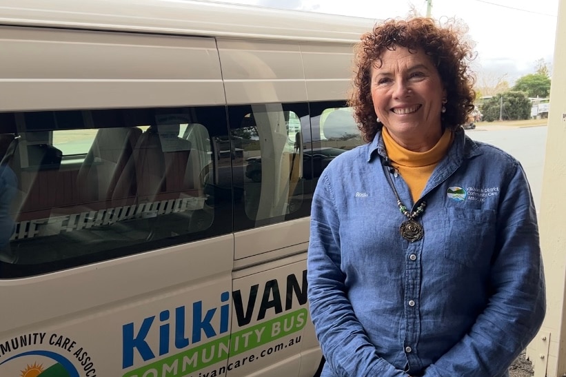A lady with curly red hair stands on the side of the road smiling in front of a white mini-van, with KilkiVan written.