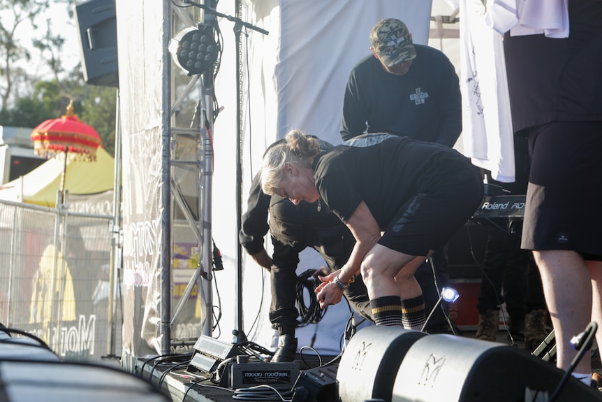 A middle-aged woman on a stage, bending over to plug in some amplifying equipment.