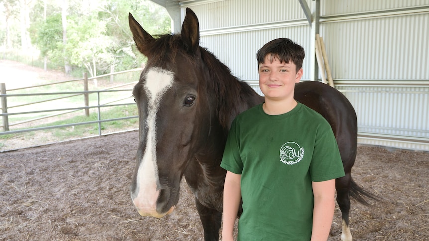 An 11 year old boy stands smiling beside a black horse with a white blaze on his face.