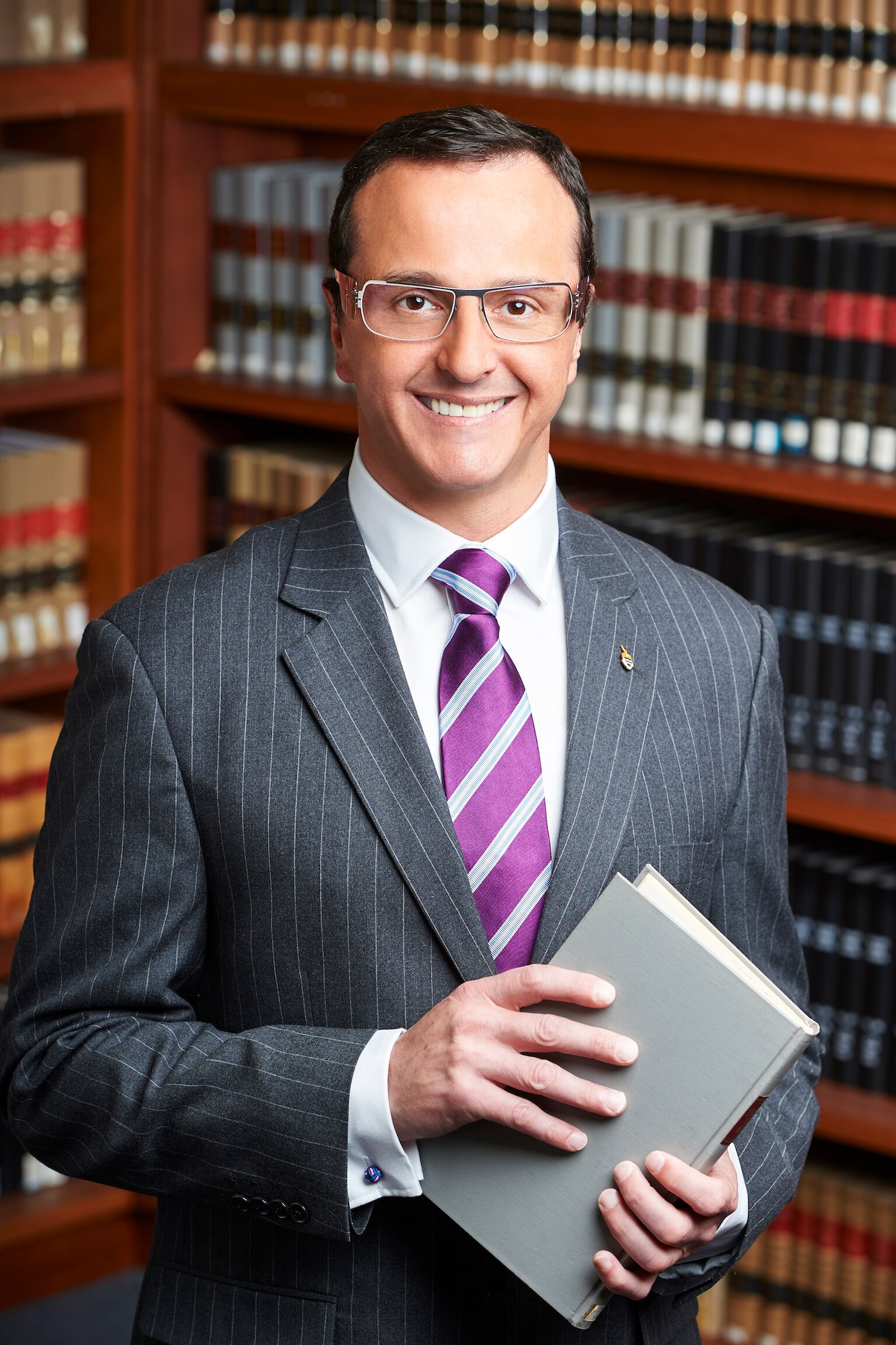 A smiling man in an expensive-looking suit, glasses, stands in front of a book case with leather-bound books.