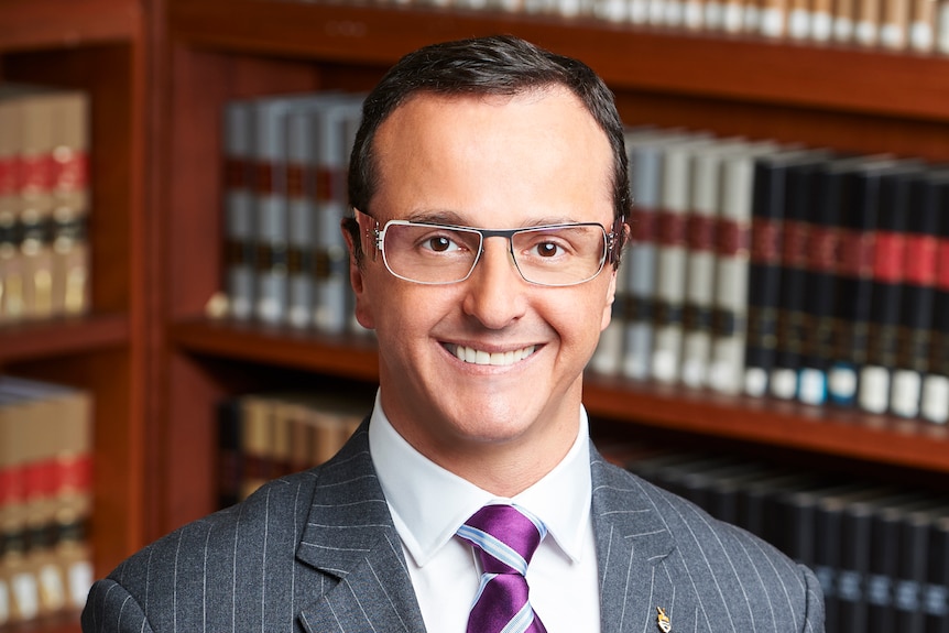 A smiling man in an expensive-looking suit, glasses, stands in front of a book case with leather-bound books.