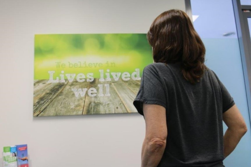 An anonymous woman with brown hair stands in front of a sign that says Lives Lived Well at the Caboolture facility.
