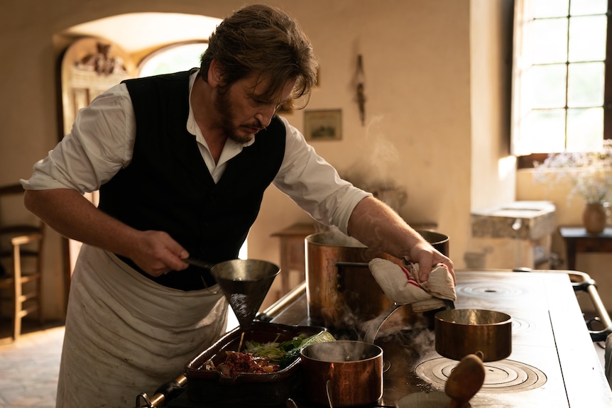 A film still of Benoît Magimel, in late 19th century dress, cooking at a stove. He's pouring flour into a saucepan.