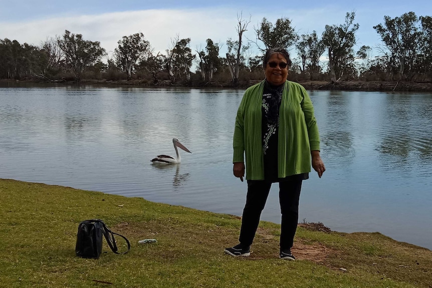 Indigenous woman, Jenni Grace, stands in front of the Murray River. She's wearing a green cardigan, a pelican is behind her.