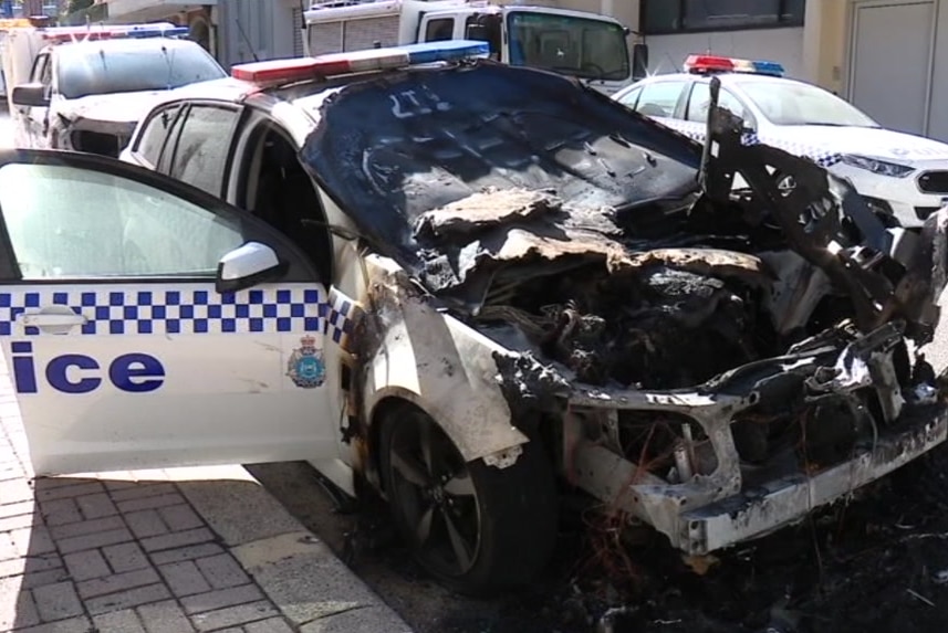 A police car sits on the side of the road with its door open after the front of the vehicle was destroyed by fire.
