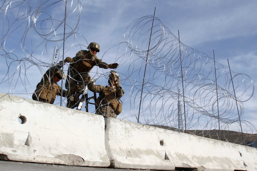 Two Marines hold another as he erects a barbed-wire fence above a concrete barricade.