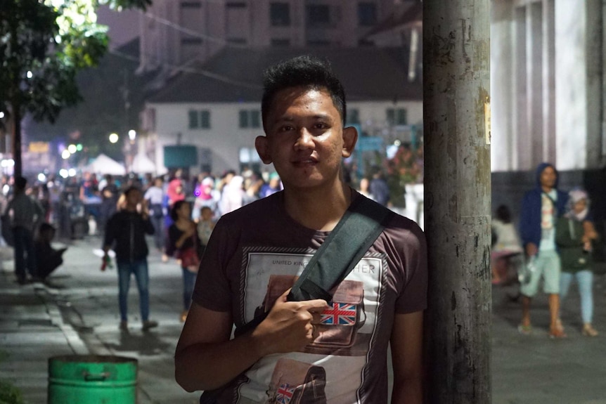 "Rangga" poses for a photo on a street while leaning against a pole.