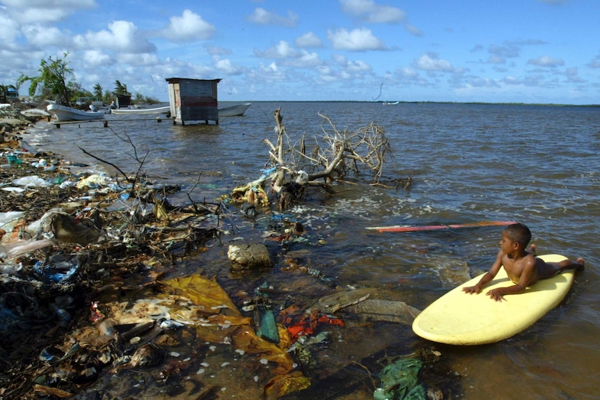 A child plays on a surfboard in a rubbish-filled lagoon
