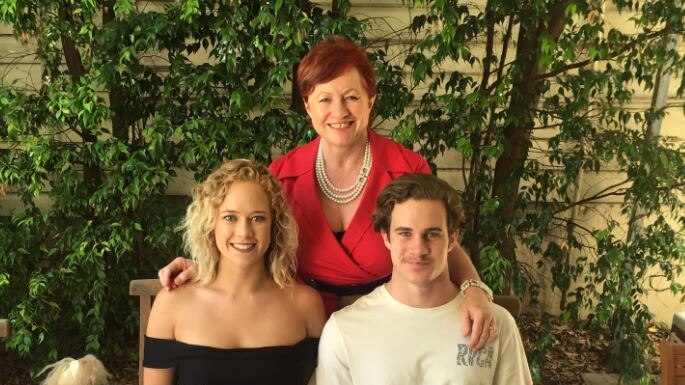 A woman with short hair stands next to two adult children, a girl with curly blonde hair and a boy with short hair