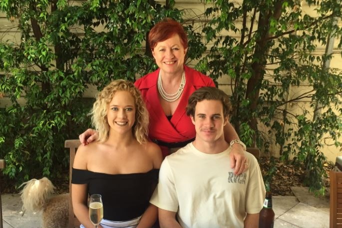 A woman with short hair stands next to two adult children, a girl with curly blonde hair and a boy with short hair