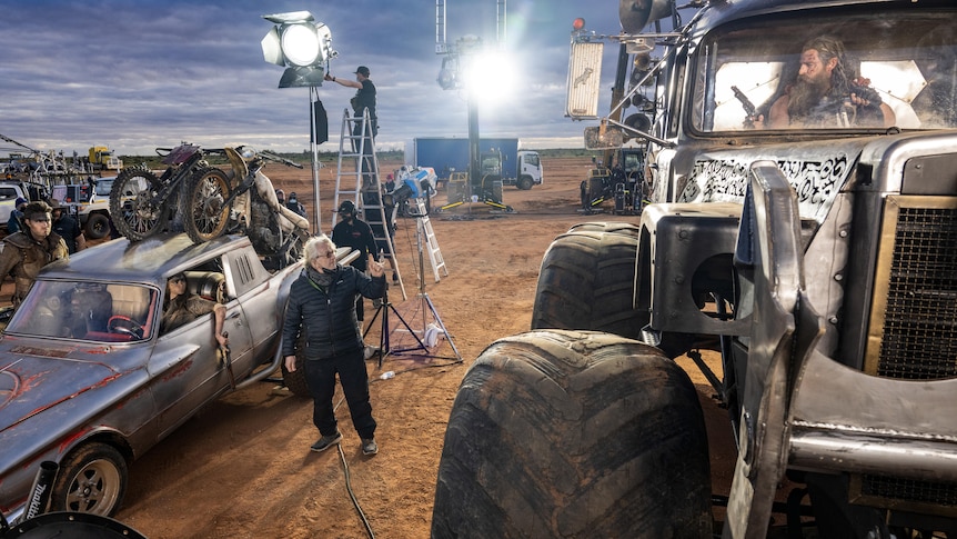 The set of Furiosa, actress Anya Taylor Joy in a small car, director George Miller standing, actor Chris Hemsworth in a big car
