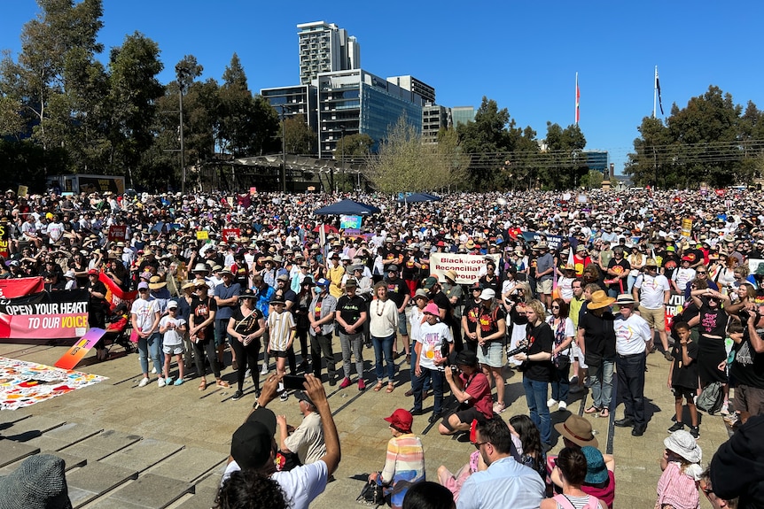 A large crowd gathered in a public square, facing a crowd, with several people holding signs in support of the Voice referendum.