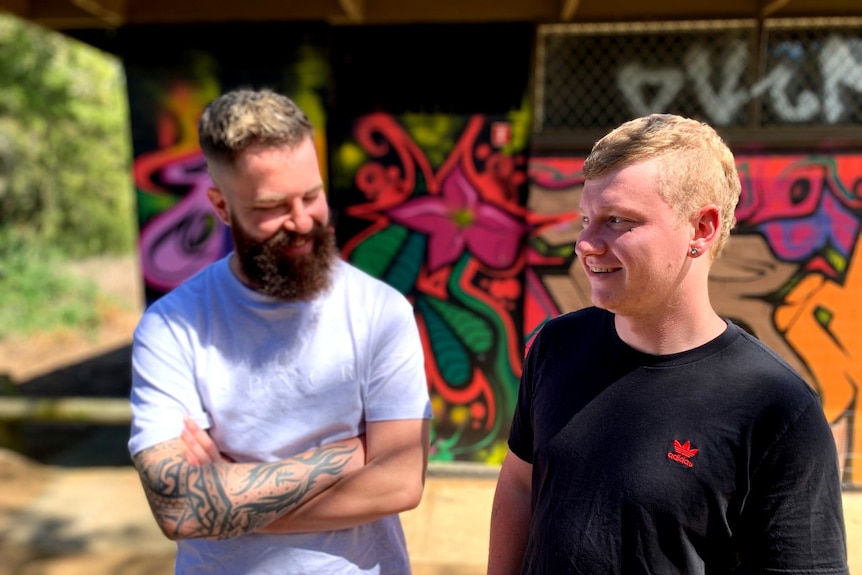 A man with a beard stands next to another young man in front of a colourful wall