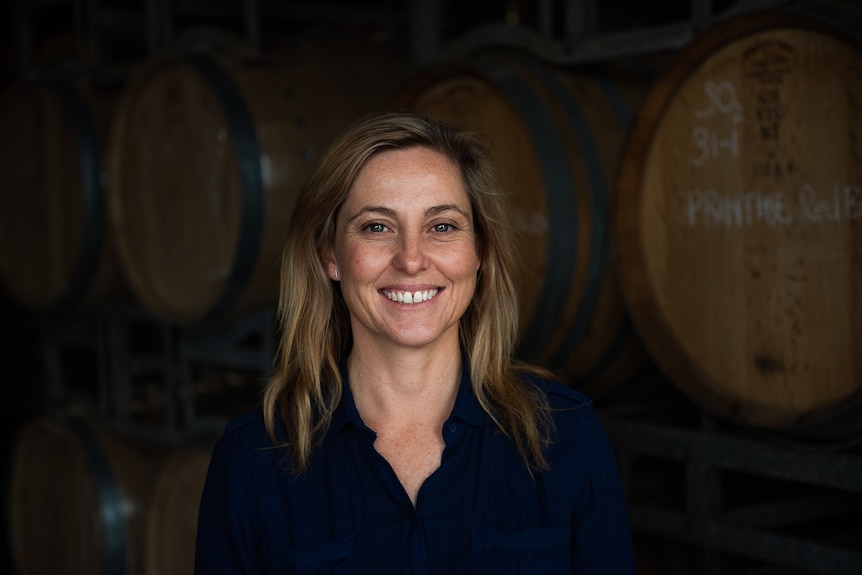 woman with long blond hair smiles wearing a black buttone shirt with wine barrels behind her