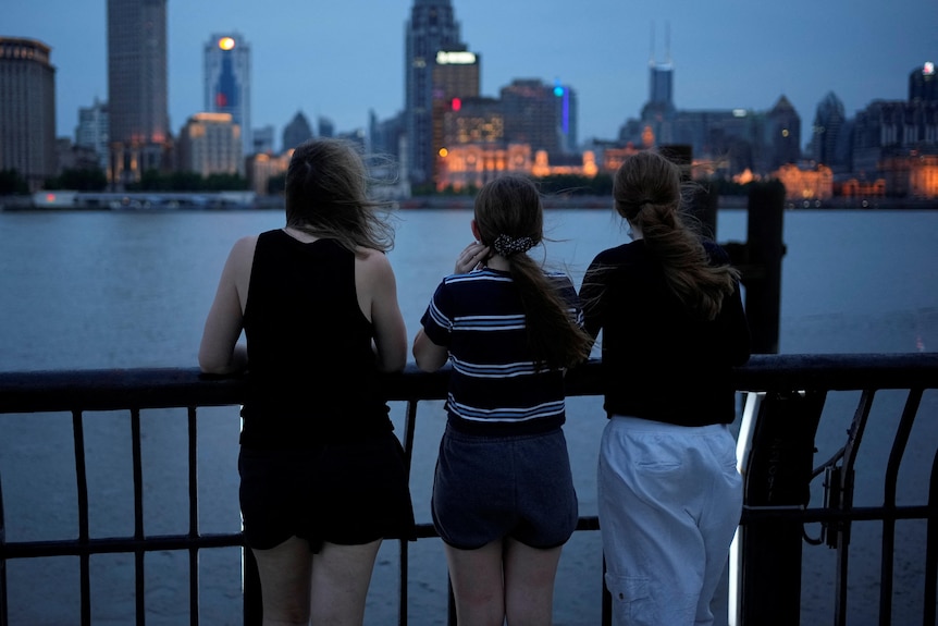 A woman stands by the water in Shanghai with her daughters in the late afternoon. The city is twinkling in the background.