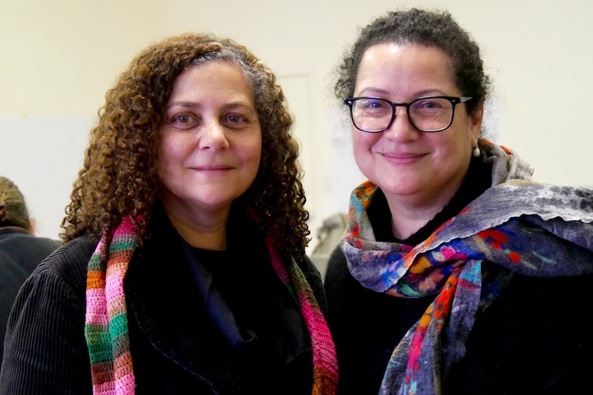 Two women smiling at the camera. The woman on the left has curly long hair and the woman on the right has glasses with a scarf  