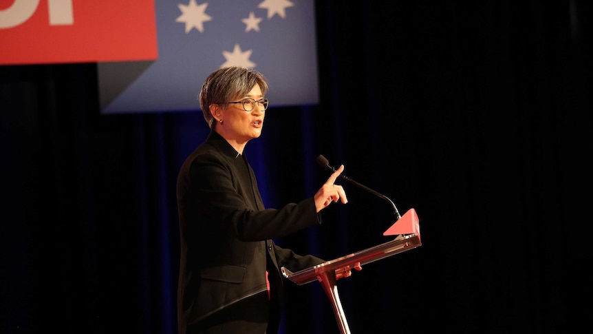 Standing at a podium holding up one finger up, Penny Wong speaks to a crowd. Labor branding hangs above her.