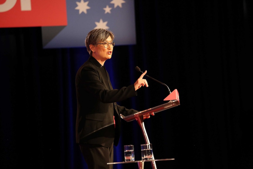 Standing at a podium holding up one finger up, Penny Wong speaks to a crowd. Labor branding hangs above her.