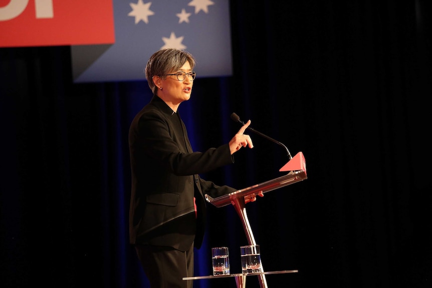 Standing at a holding one finger up, Penny Wong speaks to a crowd. Labor branding hangs above her.