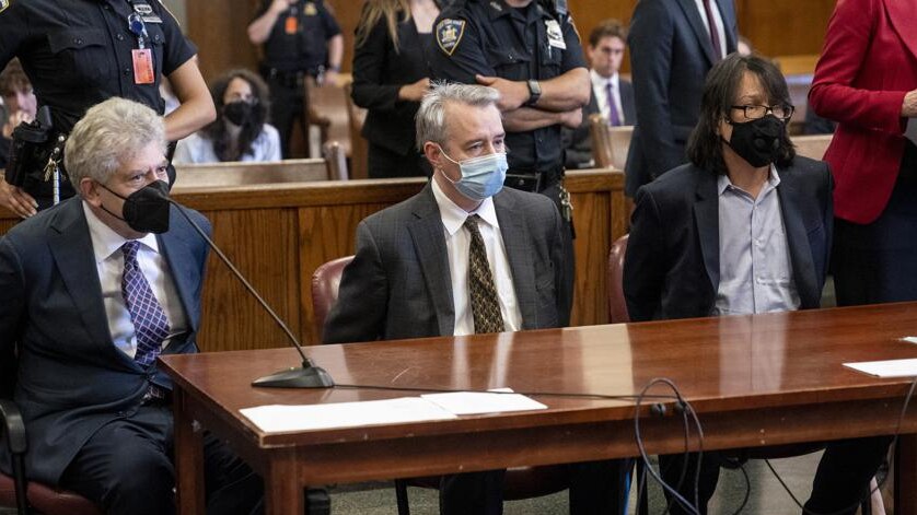 Three Caucasian men in suits wearing face masks sit in court at a long mahogany bench 