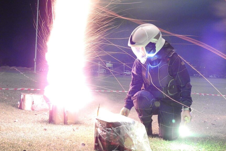 A firework shoots into the sky as a woman in a helmet and protective gear kneels to light the next firework