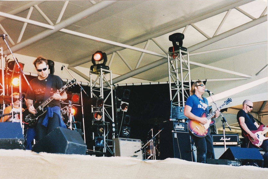 A band performs on a large stage with guitars and lighting rigs visible