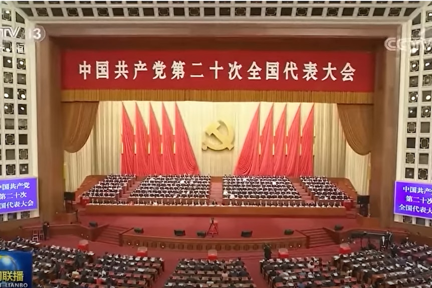 A wide angle shot of a large hall with red curtains and hammer and sickle on wall.