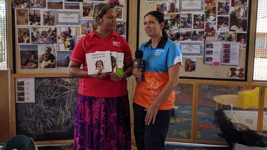 Two women stand side by side, one holding children's books