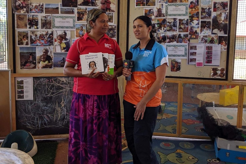 Two women stand side by side, one holding children's books