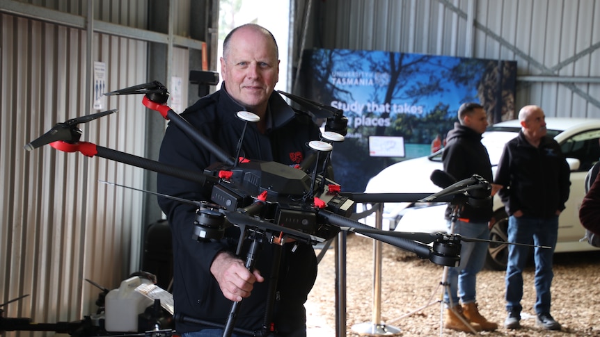 In a sprawling tin shed, Andrew holds up a big black drone, as large as an arm span.