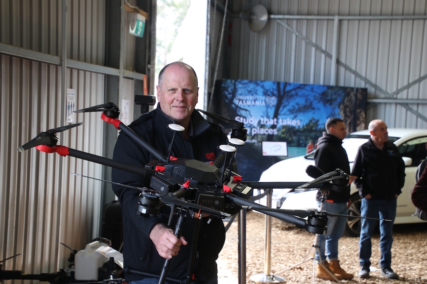 In a large tin shed, Andrew holds a large black drone, the size of an arm.