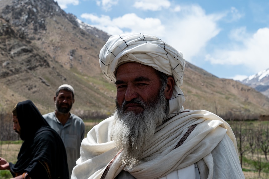 A man with a long beard and white clothes stands in front of the camera with mountains in the background