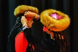 Two Uyghur women prepare for a music event. They have furry hats, braided hair, black velvet vests with bright embroidery.