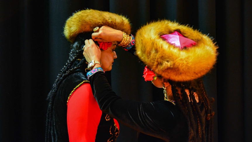 Two Uyghur women prepare for a music event. They have furry hats, braided hair, black velvet vests with bright embroidery.