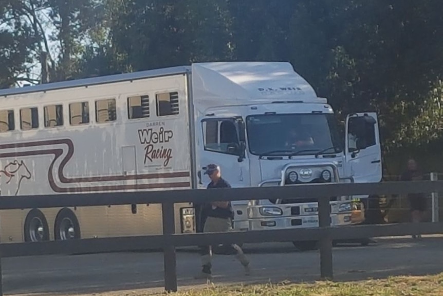 A man in a police cap walks past a large white truck with 'Weir Racing' written on the side.