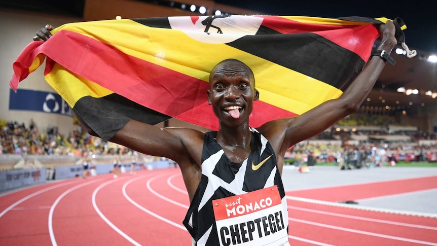 Uganda's Joshua Cheptegei sticks his tongue out as he holds up the Ugandan flag over his head after finishing his race