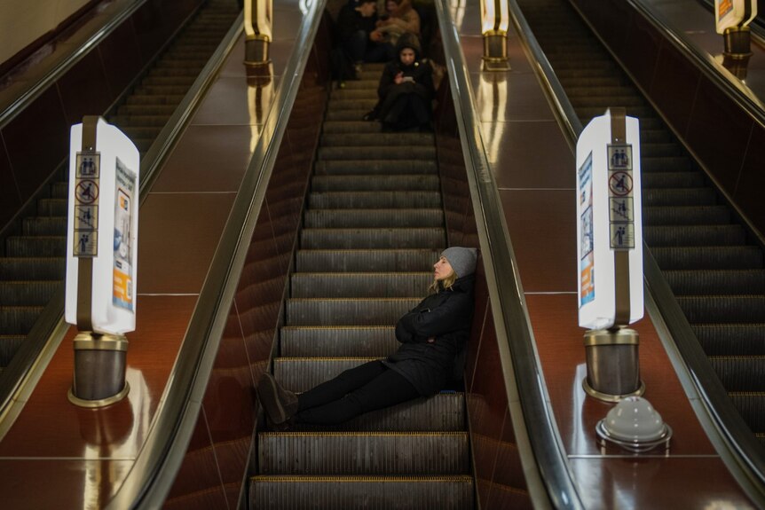A woman reclines on an inactive escalator step, others huddle on steps above her