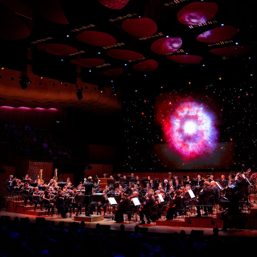 An orchestra performs on a dark stage in the Sydney Opera House. A screen behind them shows an bright magenta image from space.