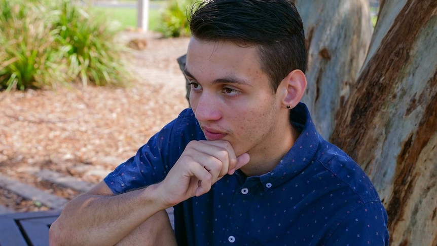 A young man sits on a park bench with his arm resting on his raised knee, looking into the distance with a neutral expression.